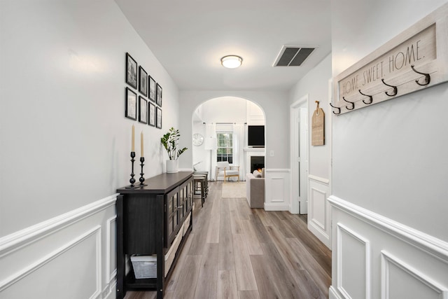 corridor featuring light wood finished floors, visible vents, arched walkways, a wainscoted wall, and a decorative wall