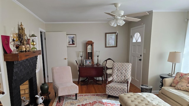 living area featuring wood finished floors, a ceiling fan, baseboards, ornamental molding, and a brick fireplace