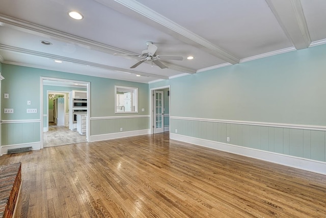 empty room with a ceiling fan, wood finished floors, a wainscoted wall, crown molding, and beamed ceiling