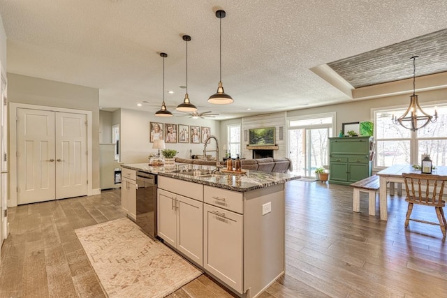 kitchen with light wood finished floors, dishwasher, an island with sink, a fireplace, and a sink