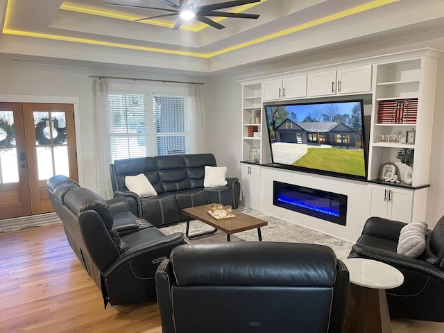 living area with a glass covered fireplace, a raised ceiling, light wood-style floors, and a ceiling fan