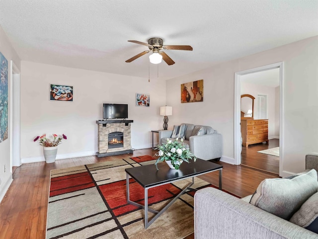 living area with hardwood / wood-style floors, a ceiling fan, baseboards, a stone fireplace, and a textured ceiling