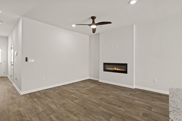 unfurnished living room featuring a glass covered fireplace, baseboards, dark wood-style flooring, and ceiling fan