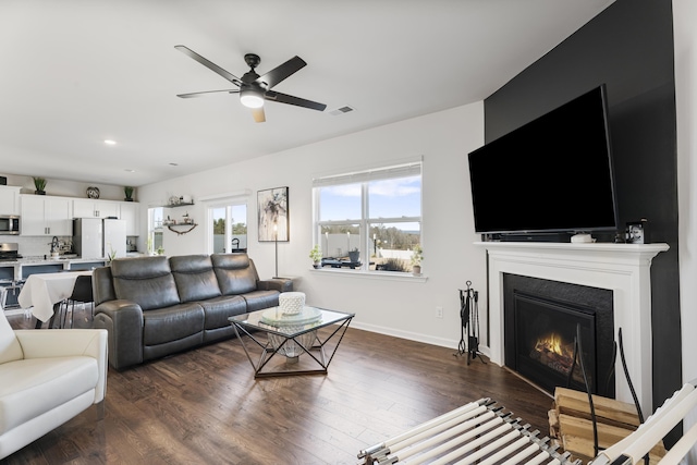 living room with a ceiling fan, baseboards, visible vents, dark wood-style flooring, and a lit fireplace