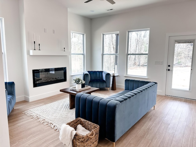 living area with visible vents, a ceiling fan, a glass covered fireplace, light wood-style floors, and baseboards