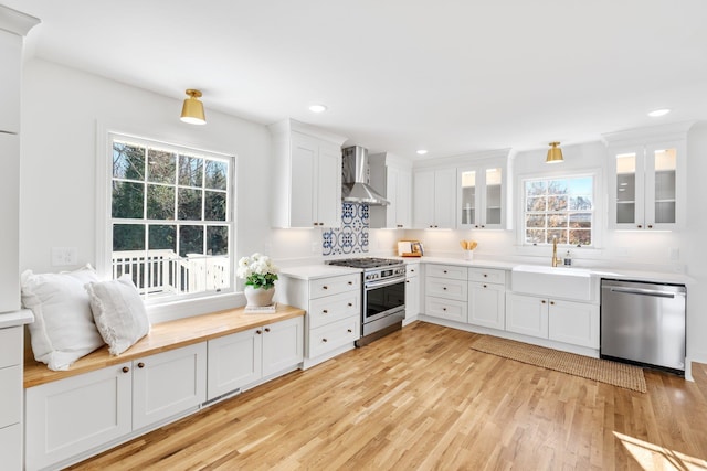 kitchen featuring appliances with stainless steel finishes, light wood-style floors, white cabinetry, wall chimney exhaust hood, and a sink