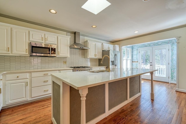 kitchen with ornamental molding, stainless steel appliances, wall chimney range hood, and a kitchen island with sink