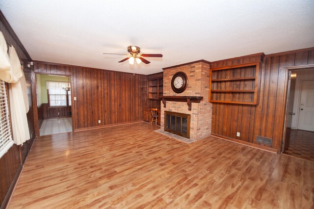 unfurnished living room with a ceiling fan, a fireplace, wooden walls, light wood-style flooring, and ornamental molding