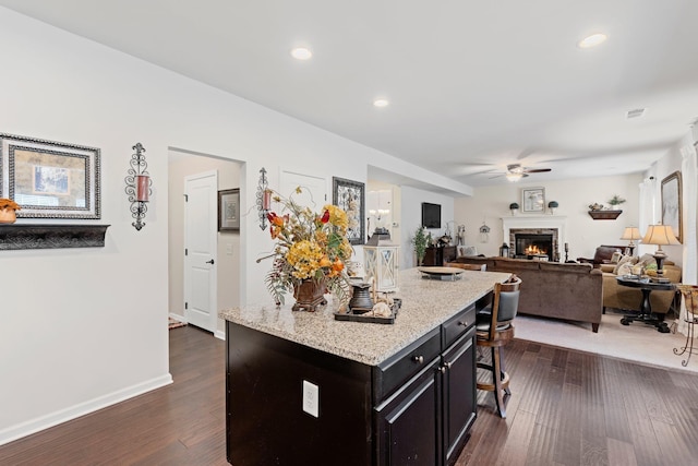 kitchen featuring ceiling fan, dark wood finished floors, a kitchen breakfast bar, a warm lit fireplace, and baseboards