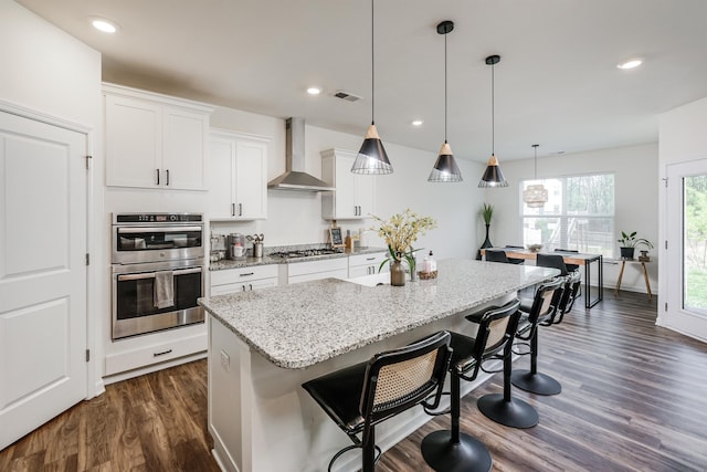 kitchen with wall chimney range hood, visible vents, stainless steel appliances, dark wood-style flooring, and a kitchen island with sink