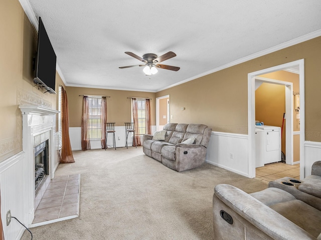 living area featuring a ceiling fan, a tiled fireplace, wainscoting, washing machine and dryer, and ornamental molding