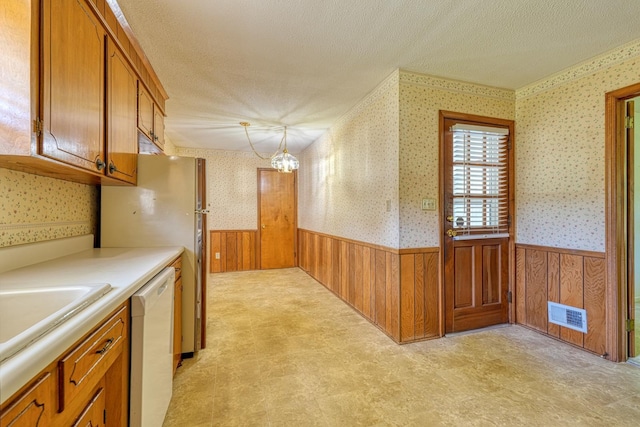 kitchen with white dishwasher, wallpapered walls, a textured ceiling, a wainscoted wall, and brown cabinets