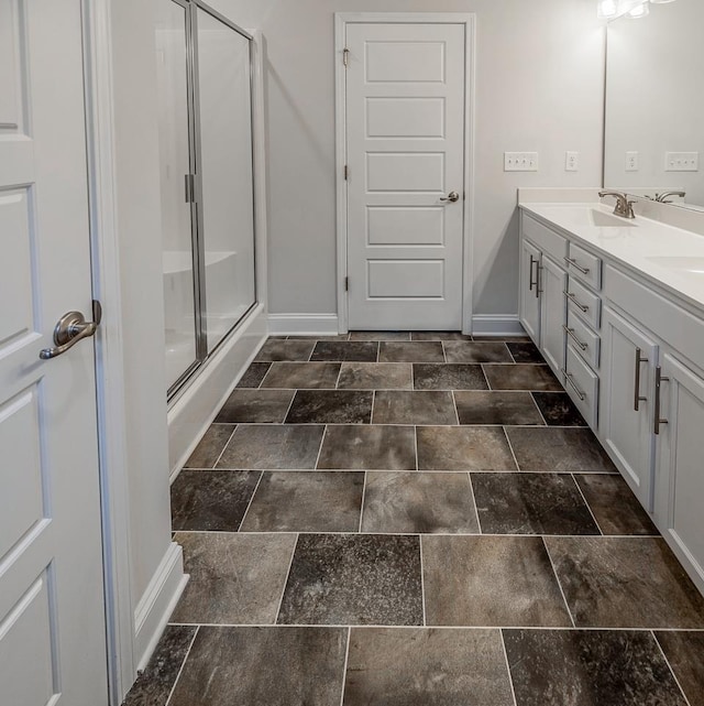 full bath featuring baseboards, a shower stall, double vanity, and stone finish flooring