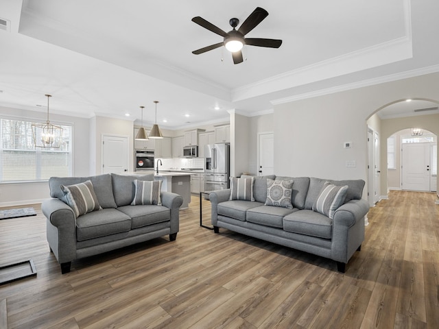 living room with a raised ceiling, arched walkways, light wood-type flooring, ceiling fan, and baseboards