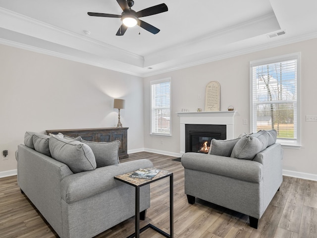 living room with a tray ceiling, a glass covered fireplace, a ceiling fan, baseboards, and wood finished floors