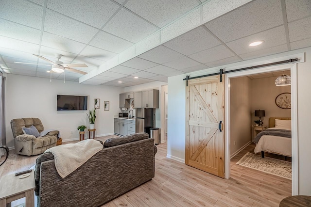 living room featuring a barn door, a drop ceiling, light wood-style floors, baseboards, and a ceiling fan