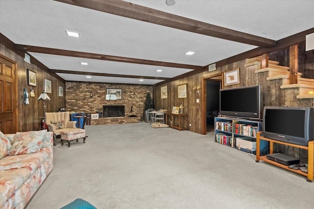 carpeted living room featuring beam ceiling, wood walls, a textured ceiling, a brick fireplace, and stairway