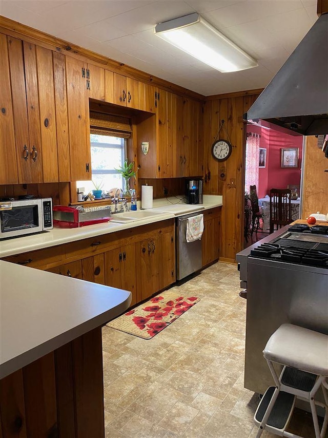 kitchen with sink, light tile floors, range hood, stainless steel appliances, and wood walls