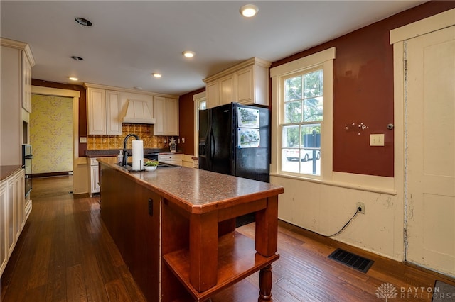 kitchen with cream cabinets, backsplash, black fridge, dark wood-type flooring, and premium range hood