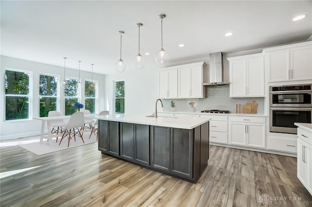 kitchen featuring light wood-type flooring, appliances with stainless steel finishes, tasteful backsplash, white cabinetry, and wall chimney range hood