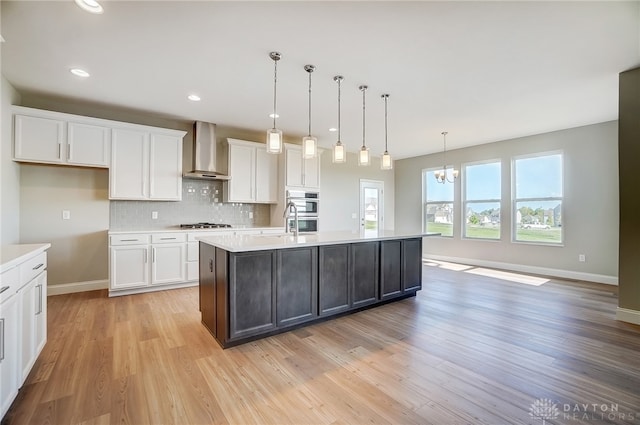 kitchen featuring decorative light fixtures, light hardwood / wood-style floors, wall chimney exhaust hood, tasteful backsplash, and a kitchen island with sink