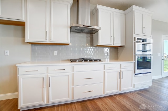 kitchen featuring appliances with stainless steel finishes, backsplash, light hardwood / wood-style floors, white cabinetry, and wall chimney range hood