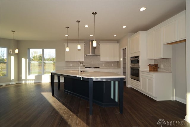 kitchen featuring an island with sink, wall chimney exhaust hood, light countertops, white cabinetry, and pendant lighting