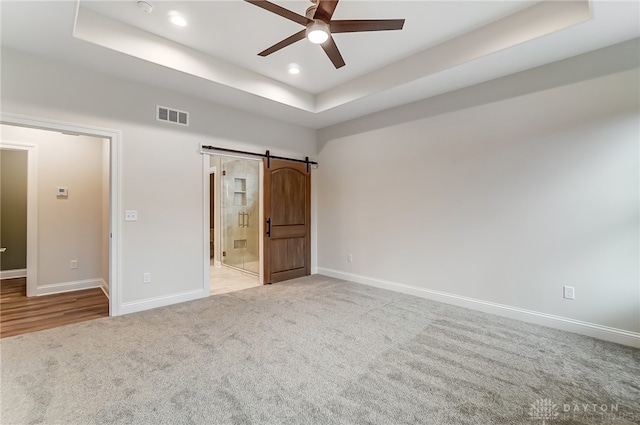 unfurnished bedroom featuring connected bathroom, ceiling fan, a raised ceiling, a barn door, and light colored carpet