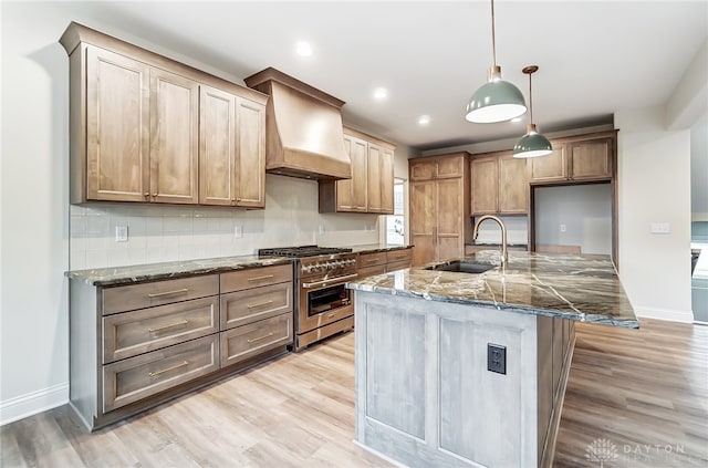 kitchen with custom exhaust hood, a kitchen island with sink, dark stone countertops, light wood-type flooring, and stainless steel range