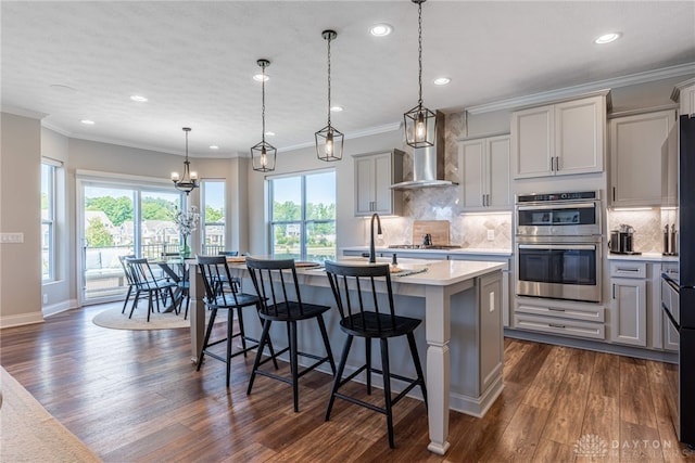 kitchen featuring gray cabinetry, a center island with sink, wall chimney range hood, decorative light fixtures, and stainless steel appliances