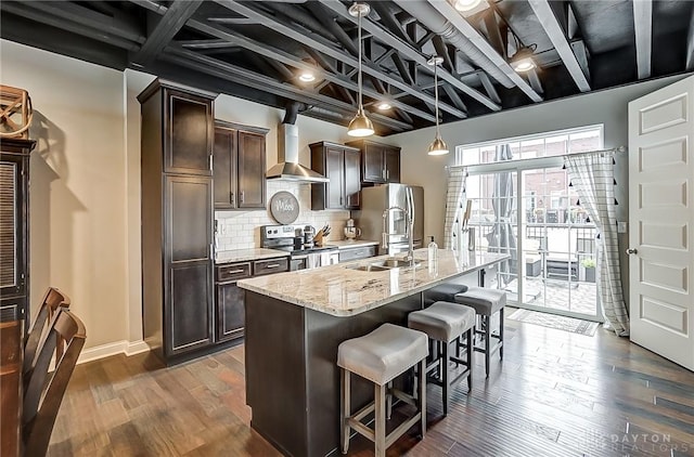 kitchen featuring wall chimney range hood, appliances with stainless steel finishes, dark brown cabinetry, an island with sink, and decorative light fixtures