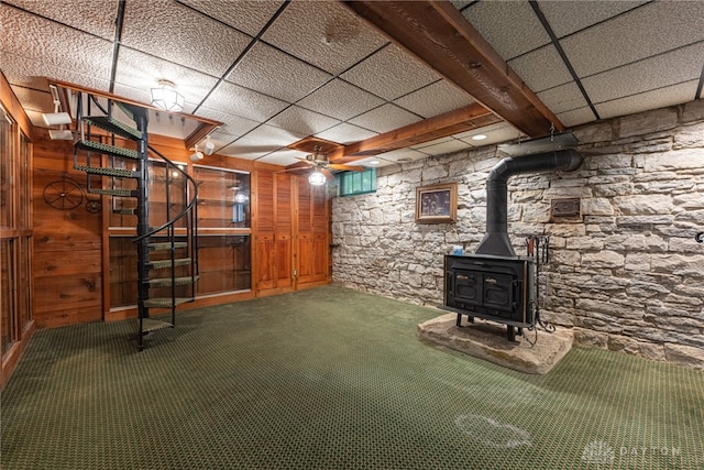 basement with ceiling fan, wooden walls, a drop ceiling, and a wood stove