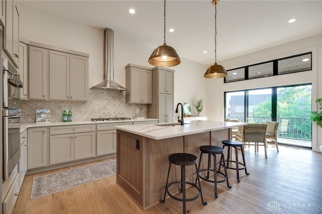 kitchen with sink, hanging light fixtures, light hardwood / wood-style floors, wall chimney exhaust hood, and a kitchen island with sink