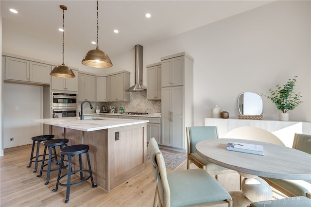 kitchen with an island with sink, light hardwood / wood-style flooring, gray cabinetry, wall chimney exhaust hood, and stainless steel appliances