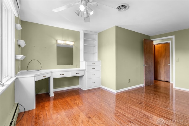 unfurnished office with built in desk, ceiling fan, a baseboard radiator, and light hardwood / wood-style floors