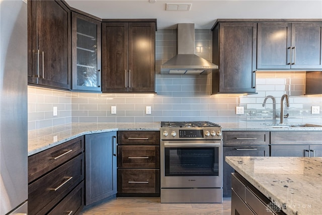 kitchen featuring wall chimney exhaust hood, light stone counters, glass insert cabinets, stainless steel appliances, and dark brown cabinets