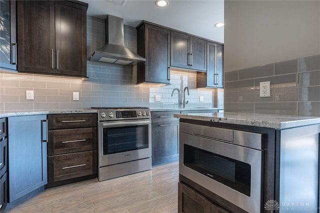 kitchen with decorative backsplash, appliances with stainless steel finishes, dark brown cabinets, light stone countertops, and wall chimney exhaust hood