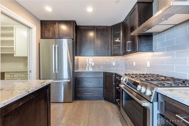 kitchen featuring wall chimney exhaust hood, light stone counters, glass insert cabinets, stainless steel appliances, and dark brown cabinets