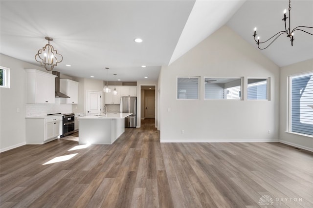 kitchen with an inviting chandelier, an island with sink, hanging light fixtures, appliances with stainless steel finishes, and wall chimney range hood