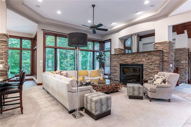 carpeted living room with a raised ceiling, ceiling fan, and a stone fireplace