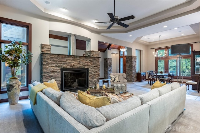 living room featuring ceiling fan with notable chandelier, a raised ceiling, and a stone fireplace