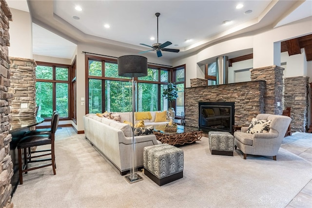 carpeted living room with ceiling fan, a stone fireplace, and a tray ceiling