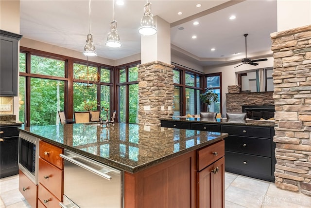 kitchen featuring hanging light fixtures, dark stone countertops, a fireplace, an island with sink, and built in microwave