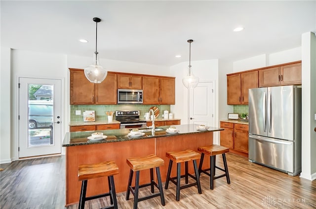 kitchen featuring stainless steel appliances, dark stone countertops, pendant lighting, light hardwood / wood-style floors, and a center island with sink