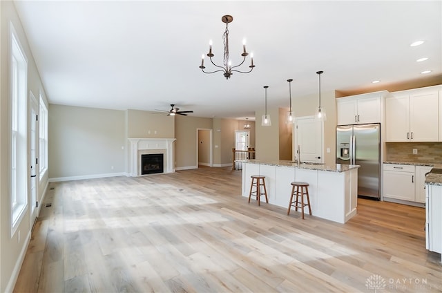 kitchen with stainless steel fridge, an island with sink, ceiling fan with notable chandelier, light wood-type flooring, and a premium fireplace