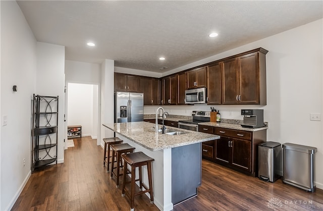 kitchen with a breakfast bar, light stone countertops, dark hardwood / wood-style flooring, stainless steel appliances, and a center island with sink