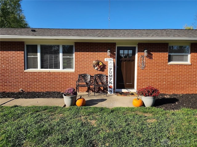 view of exterior entry featuring brick siding, roof with shingles, and a lawn