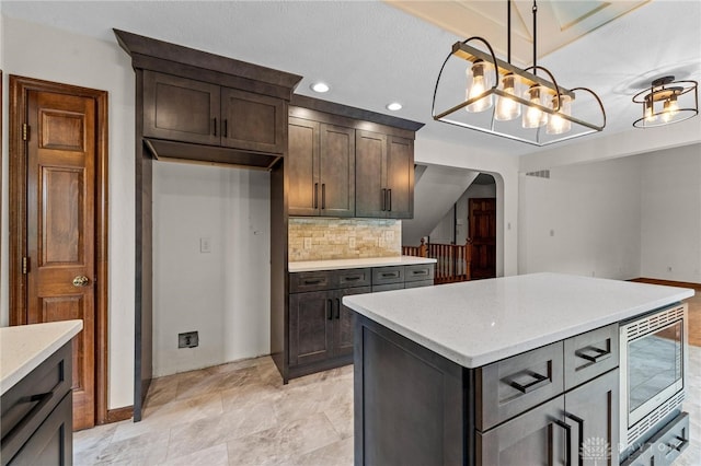 kitchen with a kitchen island, dark brown cabinetry, tasteful backsplash, stainless steel microwave, and hanging light fixtures