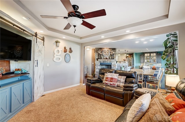 living room with ceiling fan, a raised ceiling, a barn door, a stone fireplace, and light colored carpet