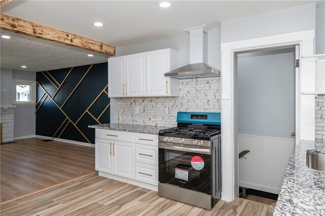 kitchen with light wood-type flooring, stainless steel range with gas stovetop, wall chimney exhaust hood, white cabinets, and beam ceiling